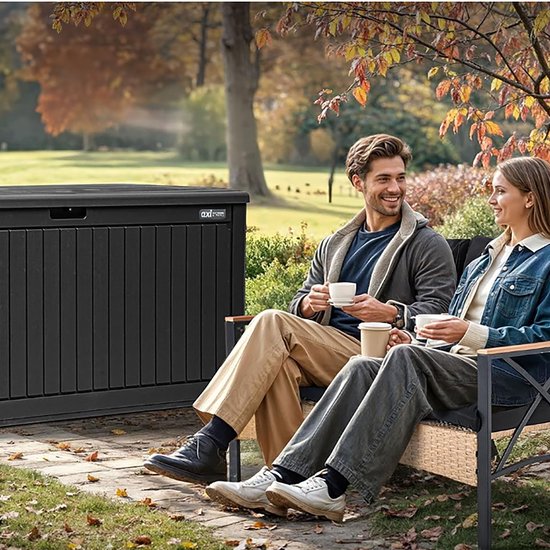 Zwarte kunststof tuinopbergbox in een tuinsetting, met een man en vrouw die koffie drinken op een bankje ernaast.