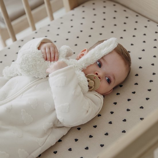 Baby met speen en knuffel in een ledikantje met zandkleurig hoeslaken met zwarte hartjes.