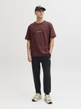 Man in een donkerrode T-shirt en zwarte joggingbroek met witte 'Jack & Jones' opdruk, zwarte pet.