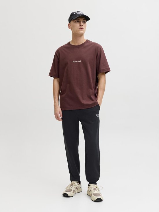 Man in een donkerrode T-shirt en zwarte joggingbroek met witte 'Jack & Jones' opdruk, zwarte pet.