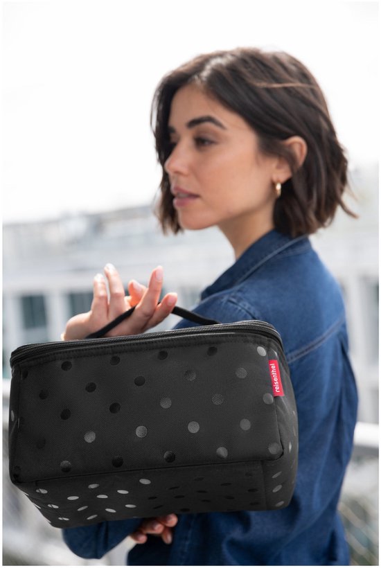 Vrouw met sproeten, gekleed in een spijkerblouse, draagt een zwarte toilettas met stippen.