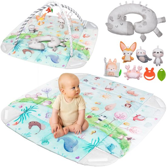 Een baby zit op een speelmat met boog en speeltjes. De mat heeft een waterdiertjes print. Naast de mat liggen losse speeltjes en een baby.