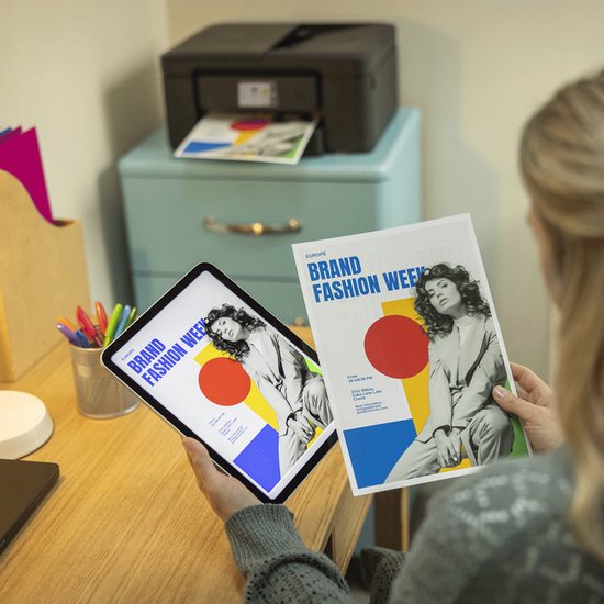 Vrouw met tablet en geprinte flyer 'BRAND FASHION WEEK' naast zwarte printer.