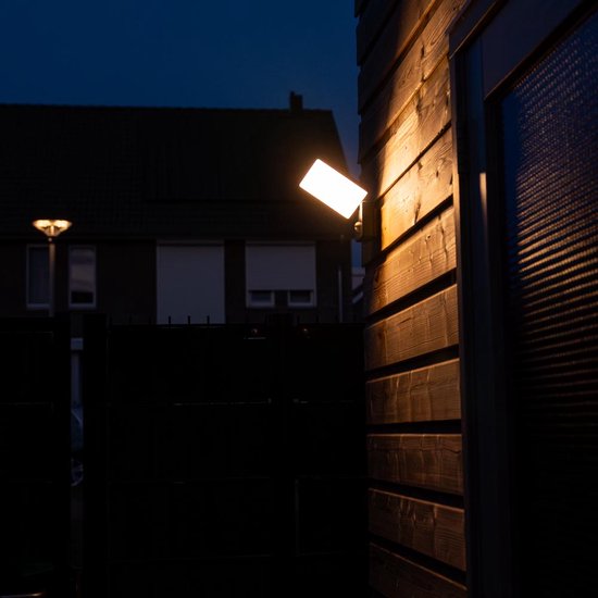 Zwarte wandlamp met sensor op houten schutting, verlicht de omgeving in de schemering.
