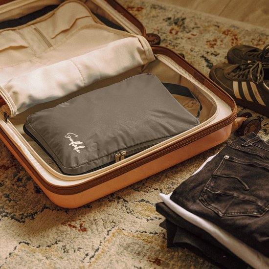 Grijze packing cube met rits in open koffer, naast jeans en sneakers.