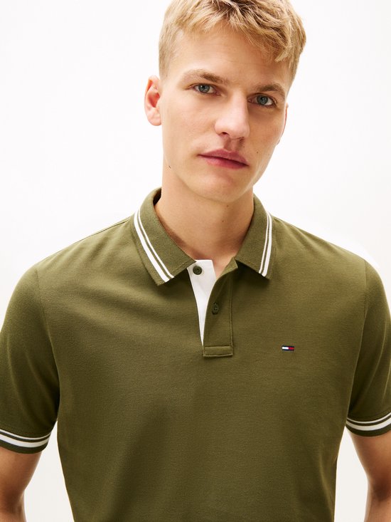 Een man met kort blond haar draagt een olijfgroene polo met witte strepen op de kraag en mouwen.