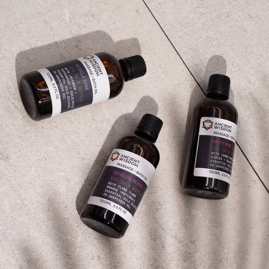 Drie amberkleurige flesjes massageolie met zwarte dopjes op een grijze tegelvloer. Van links naar rechts: 'MUSCLE EASE', 'SENSUAL BLEND', 'SPICY RUB'.