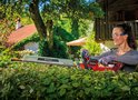 Een vrouw met veiligheidsbril snoeit een heg met een rode Einhell heggenschaar, met een huis op de achtergrond.