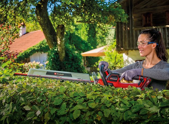 Een vrouw met veiligheidsbril snoeit een heg met een rode Einhell heggenschaar, met een huis op de achtergrond.