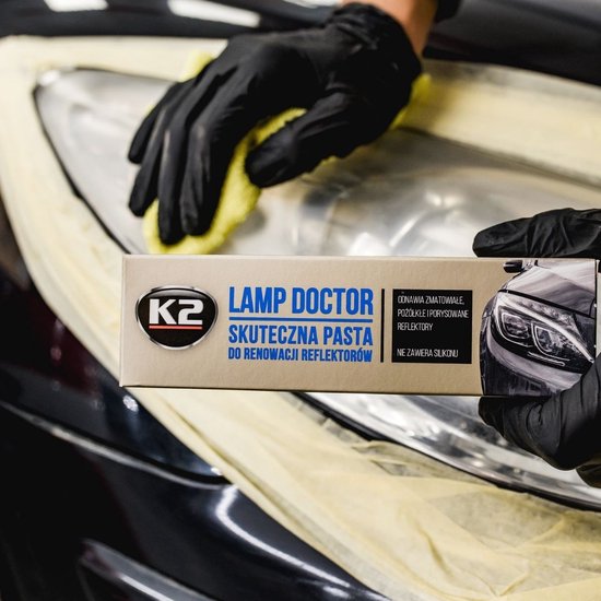 Hand met zwarte handschoen houdt K2 Lamp Doctor doos boven auto koplamp.