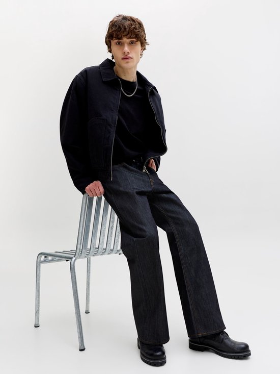 Model zittend op een stoel, gekleed in een zwarte jas en donkere jeans met wijd uitlopende pijpen.