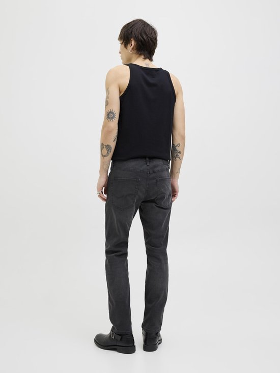 Man met tatoeages, staand met de rug naar de camera, gekleed in een zwarte tanktop, grijze spijkerbroek en zwarte laarzen.