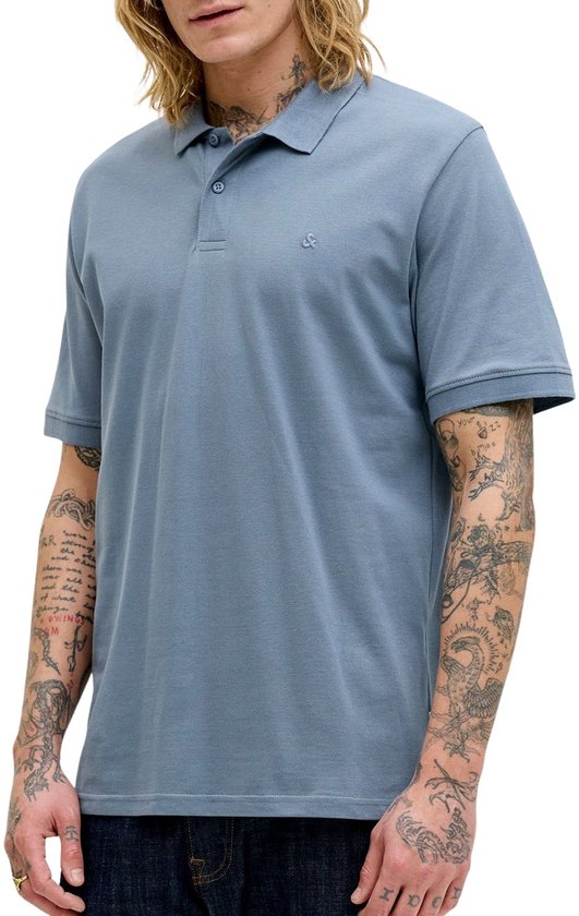 Man met lang blond haar draagt een blauwgrijze polo. Volledige body shot, met tatoeages op armen.