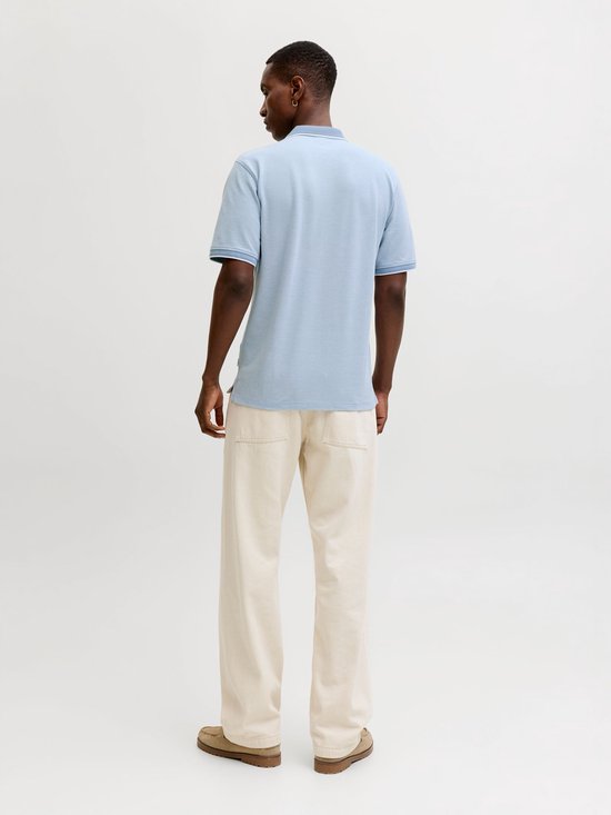 Man in lichtblauw poloshirt met witte kraag en manchetten, beige broek en bruine schoenen.