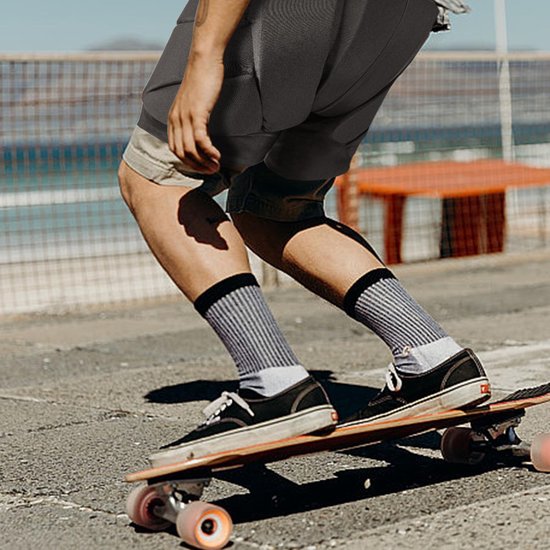 Een persoon op een skateboard, draagt een grijze short, met padding op de heupen, grijze sokken met strepen en zwarte sneakers.