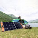 Zwart Jackery zonnepaneel en oranje Jackery solar generator op gras bij een meer, man met boot op achtergrond.