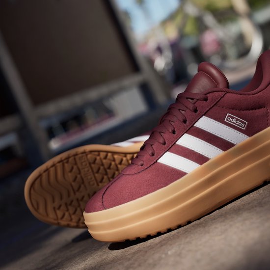 Bordeauxrode Adidas sneakers met witte strepen, dikke gum-zolen, op de grond.