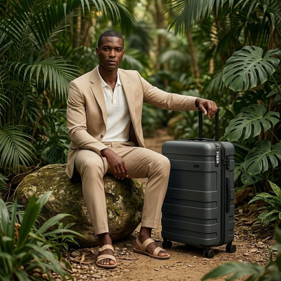 Man in beige pak met witte polo en sandalen, zittend op een rots met een donkergroene koffer in de jungle.