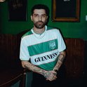 Man met baard en tatoeages, draagt een wit met groen gestreept voetbalshirt met 'Guinness' opdruk.
