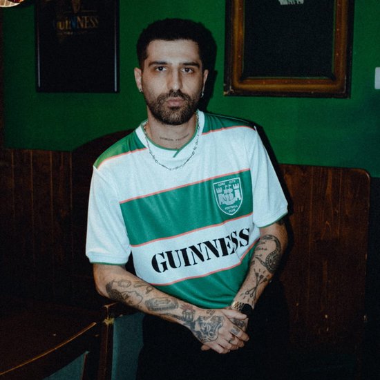 Man met baard en tatoeages, draagt een wit met groen gestreept voetbalshirt met 'Guinness' opdruk.