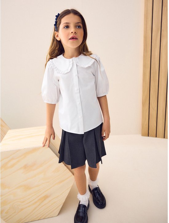 Meisje draagt witte blouse met rucheskraag, grijze rok, witte sokken en zwarte schoenen.