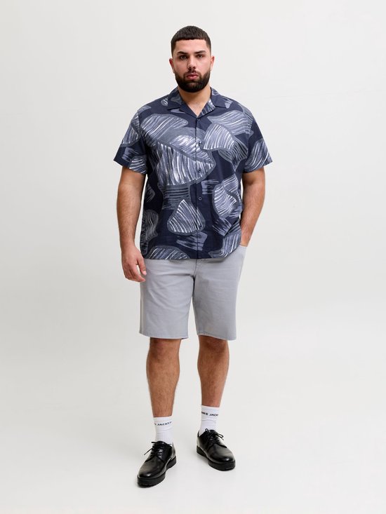 Model in een blauw, kort overhemd met een grijze abstracte bladprint, gecombineerd met een grijze korte broek en witte sokken.