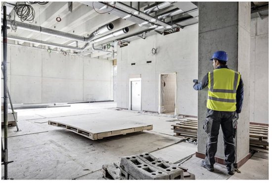 Bouwvakker met geel hesje en blauwe helm meet met afstandsmeter in ruimte met pallets.