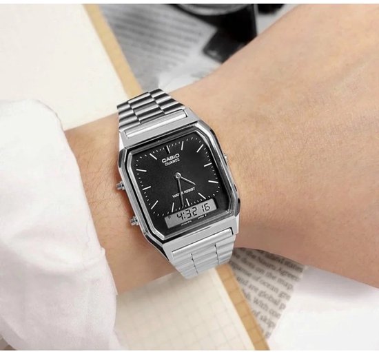 Porteuse montrant une montre argentée avec bracelet métallique, cadran noir, heure analogique et numérique, portée avec une blouse blanche.