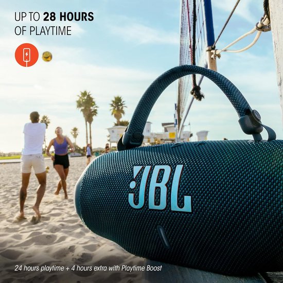 Enceinte JBL noire sur la plage, avec deux personnes à l'arrière-plan.