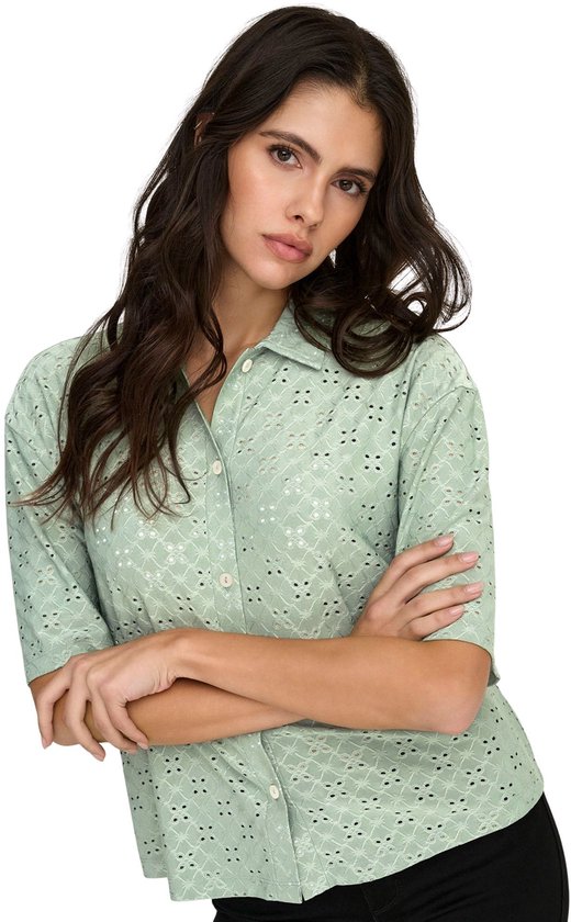 Model met gekruiste armen in een lichtgroene blouse met broderie anglaise, korte mouwen en knopen.