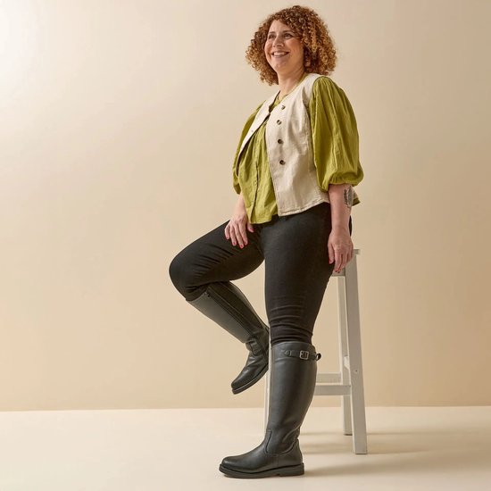 Vrouw met krullend haar, gekleed in een groene blouse en een beige vestje, draagt zwarte laarzen.