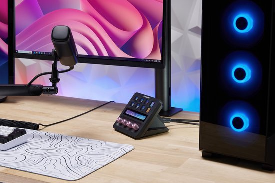 Elgato Stream Deck + op een zwarte standaard, naast een monitor en een gaming PC met blauwe verlichting.