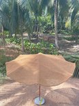 Een grote, ronde parasol van natuurlijke kokosvezel met een golvend rand staat op de grond in een tropische tuin.