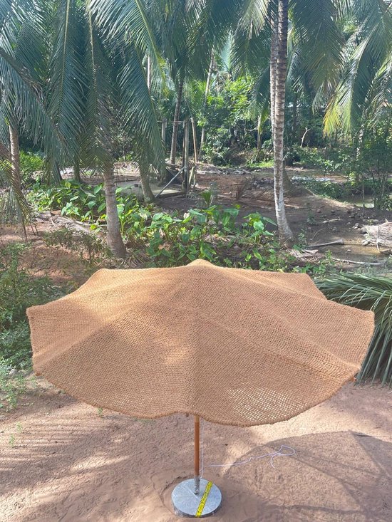 Een grote, ronde parasol van natuurlijke kokosvezel met een golvend rand staat op de grond in een tropische tuin.