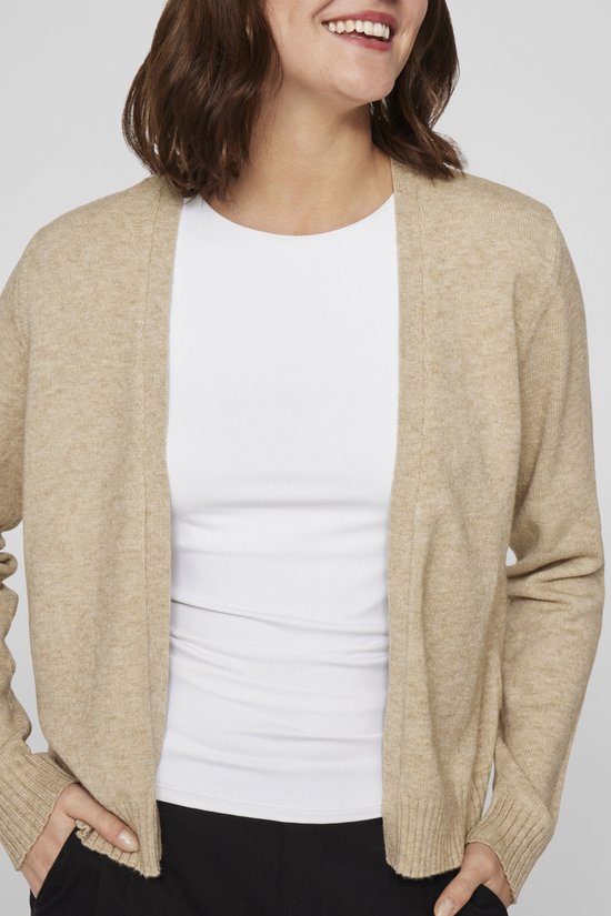 Detail van een vrouw met haar handen in haar zakken, draagt een beige vest over een wit shirt.
