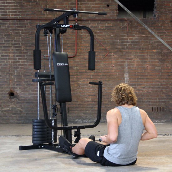 Man met krullend haar, grijs T-shirt en zwarte korte broek zit aan het Focus Fitness Unit 2 krachtstation.