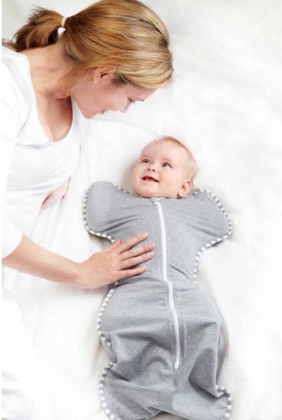 Een baby glimlacht naar een vrouw die haar met haar hand aanraakt, terwijl de baby in een grijze Swaddle UP slaapzak ligt.