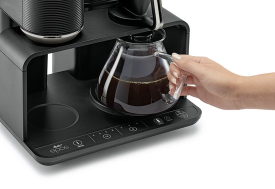Een hand pakt een glazen koffiekan uit een zwart koffiezetapparaat met digitaal bedieningspaneel.