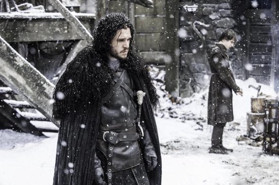 Een man met lang zwart haar en een zwarte, leren outfit met een grote, harige zwarte cape, staat in de sneeuw.