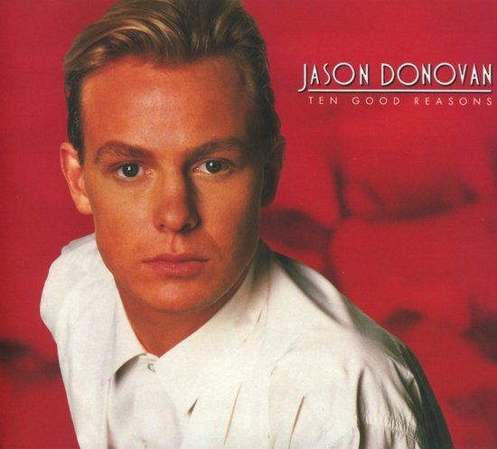 Close-up van Jason Donovan met blonde kuif, wit overhemd, tegen rode achtergrond.
