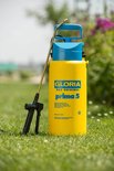 Gele Gloria Prima 5 drukspuit met blauwe handgreep op gras, met bloemen op de achtergrond.