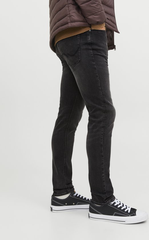 Zijaanzicht van een man in donkerbruine pufferjas, bruine trui en donkergrijze slim fit jeans met witte sneakers.
