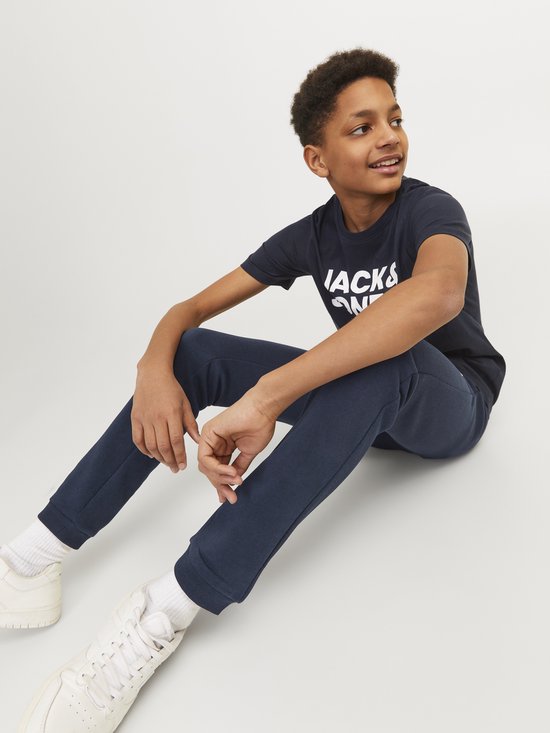 Jongen zittend in een marineblauwe joggingbroek en T-shirt met 'JACK & JONES' opdruk, met witte sneakers.