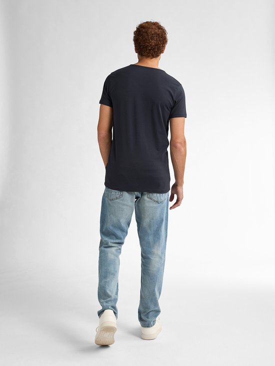 Achterkant van een man met krullend bruin haar, in een donkerblauw V-hals T-shirt, spijkerbroek en witte sneakers.