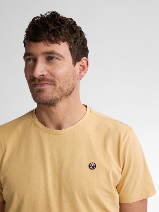Man met licht getinte huid draagt een donkergeel T-shirt met een klein rond logo met 'P' op de linkerborst.