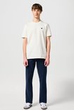 Man in een crème T-shirt met klein logo en een donkerblauwe spijkerbroek.