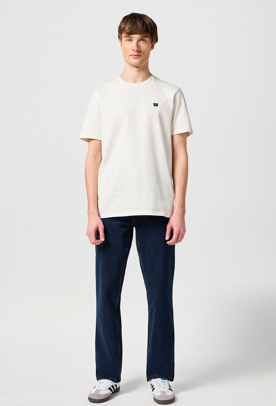 Man in een crème T-shirt met klein logo en een donkerblauwe spijkerbroek.