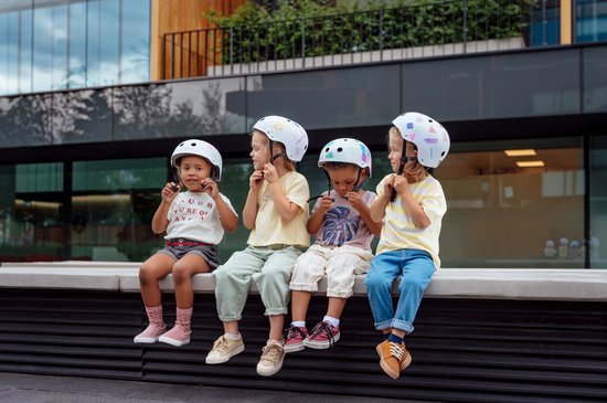 Vier kinderen met helmen zitten op een rand, twee hebben witte helmen, twee met opdruk.
