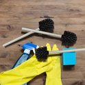 Twee toiletborstels met roestvrijstalen handvat, gele handschoenen, blauwe spuitfles en spons op houten achtergrond.