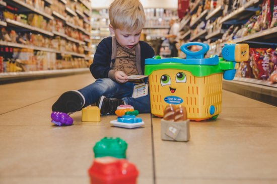 Jongen in winkel, zittend op de grond, speelt met een speelgoedwinkelwagen en boodschappen.